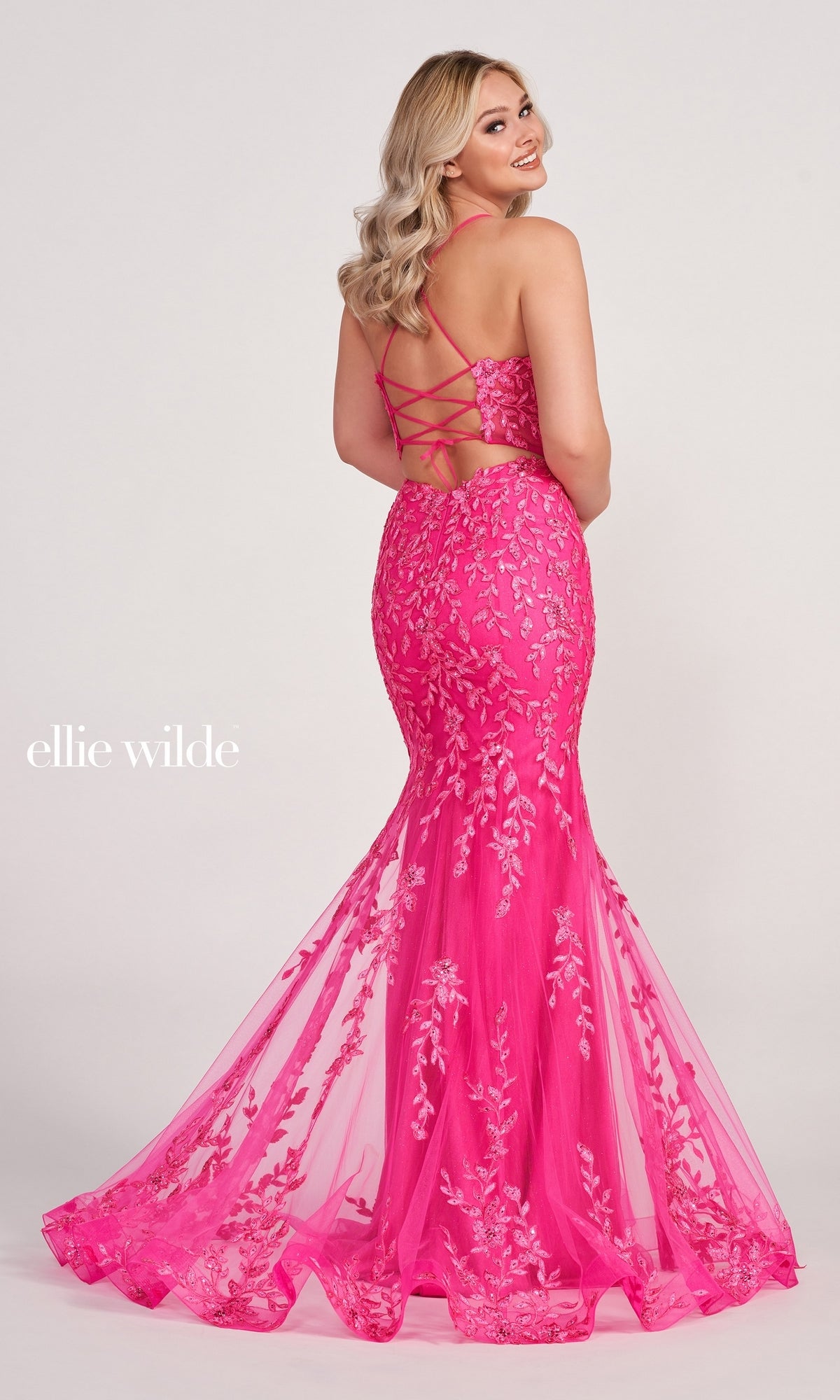 A woman wearing the Lace Applique Over Glitter Jersey Gown EW34090 in bright pink, featuring a mermaid silhouette, embroidered lace appliqué, and a lace-up open back, stands looking over her shoulder.